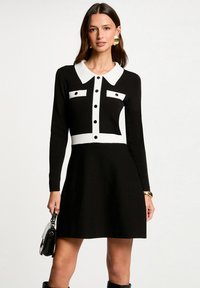 Vestido negro con cuello y detalles blancos, mangas largas, botones decorativos, forma de falda acampanada, tejido texturizado, combinado con un bolso.