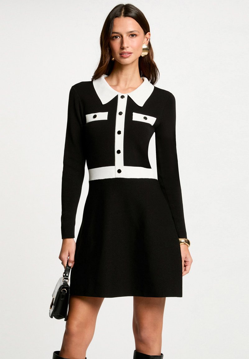 Vestido negro con cuello y detalles blancos, mangas largas, botones decorativos, forma de falda acampanada, tejido texturizado, combinado con un bolso.