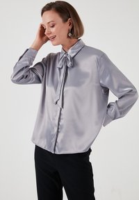 Light gray satin shirt with a bow tie at the collar, long sleeves, and a slightly loose fit, paired with dark trousers.