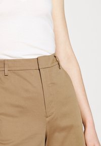 Un pantalon en coton beige clair présente une taille cintrée, des poches latérales et une coupe droite, le tout complété par une texture lisse et mate.