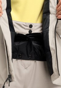 Veste grise clair avec une doublure intérieure noire, dotée de deux boutons noirs et d'une taille élastique. Fini lisse avec une fermeture éclair.