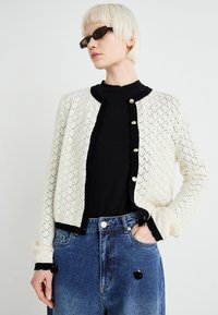 Cardigan tricotat în crem cu un design din dantelă și margini contrastante negre. Prezintă un guler rotund și nasturi aurii. Asortat cu blugi cu talie înaltă.
