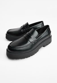 Zwarte leren loafers met stevige loopzolen, een gladde afwerking en een banddetail over de wreef, tentoongesteld op een witte achtergrond.
