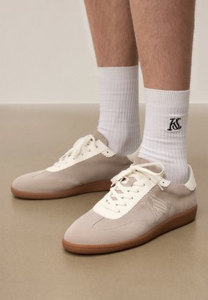 Beige ja valge seemisnahast tossude paar valgete paeltega, kantud valgete soonikkoes sokkidega, millel on must "KS" logo pahkluu kohal.