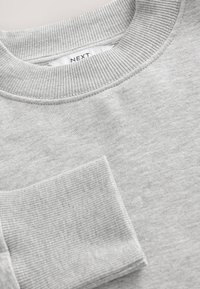 Pilkas sportinis megztinis su ribiniu apykakliu ir rankogaliu. Minkštos audinio tekstūros, apvalus iškirptė ir ma ža kišenės detalė priekyje. Matomas prekės ženklo žymėjimas.