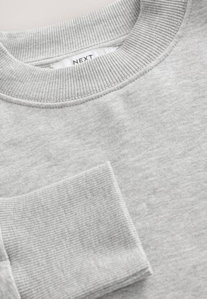 Šedá mikina s žebrovaným límcem a manžetami. Měkká textura látky, kulatý výstřih a malá kapsa vpředu. Viditelná značka produktu.