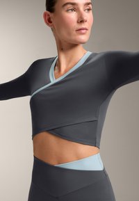 Top sportivo a maniche lunghe grigio con design incrociato, caratterizzato da scollo e bordi azzurri chiari. Tessuto liscio, silhouette aderente, con dettagli ritagliati.