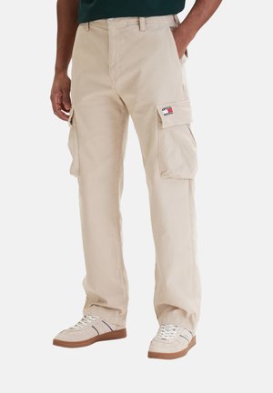 Pantalones cargo beige con grandes bolsillos laterales y parche con logo en el bolsillo derecho, combinados con zapatillas beige y camiseta verde oscuro.