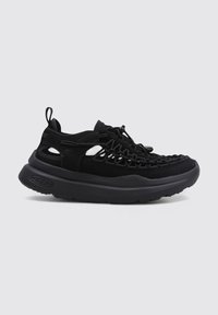 Zapatillas deportivas negras que cuentan con una parte superior de textil, un sistema de cordones trenzados, una lengüeta para tirar y una suela de goma gruesa con patrones texturizados.