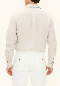 Camicia di lino grigio chiaro con piega sul retro, maniche lunghe e polsini abbottonati, abbinata a pantaloni bianchi con tasche posteriori e dettagli in metallo.