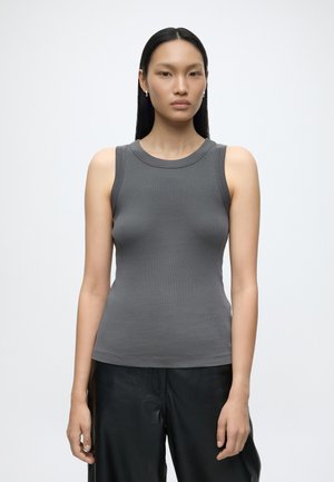 Femme aux cheveux noirs et raides, portant un haut gris ajusté sans manches et un pantalon noir, debout devant un fond blanc uni.