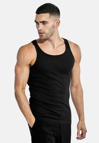Black ribbed tank top with wide straps, fitted design, and soft texture, paired with black pants. Model stands with one hand in pocket.