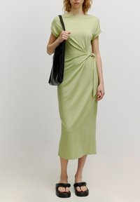 Knielanges, blassgrünes Kleid mit kurzen Ärmeln und einer Knoten-Optik an der Taille. Getragen mit einer schwarzen Schultertasche und klobigen schwarzen Sandalen.