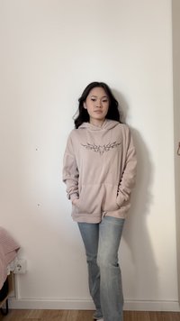 Jauna sieviete ar gariem tumšiem matiem, nēsājot bēša kapuci ar melnu cilšu dizainu un gaiši zilas džinsas, stāv pret vienkāršu sienu.