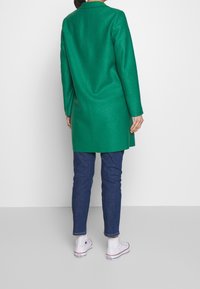 Abrigo de lana verde con un diseño entallado, mangas largas y corte recto, combinado con vaqueros azules y zapatillas blancas. Textura suave.