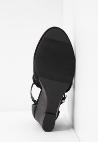 Sandale compensée noire avec talon texturé et semelle en caoutchouc, taille 4, positionnée semelle vers le haut contre un fond blanc d'angle.