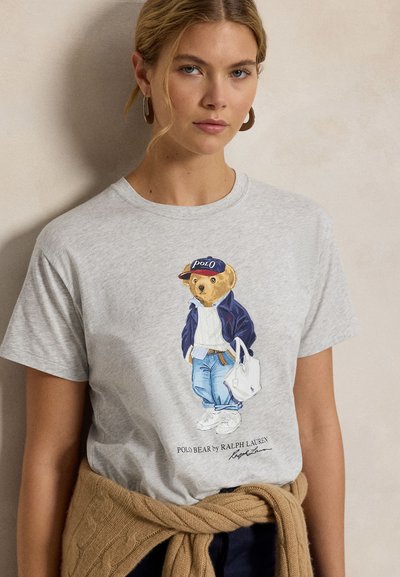 Mladá žena nosící šedé tričko s grafikou medvěda v běžném oblečení a držícího bílou tašku.