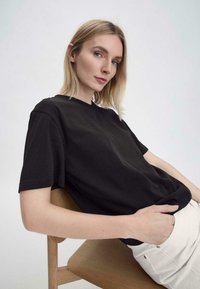 Schwarzes Baumwoll-T-Shirt mit kurzen Ärmeln, Rundhalsausschnitt und lockerer Passform, kombiniert mit weißen, hochtaillierten Hosen und sitzt auf einem Holzstuhl.