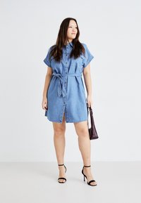 Denim skjortekjole med korte ermer, knapfront, belted midje og knelangt fald. Kombinert med svarte stroppheels og en burgunder håndveske.