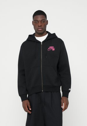 Mlad moški nosi črn zadrgo Nike SB hoodie s pisanjem v roza barvi, črne hlače in belo majico, stoji ob enotni svetli ozadju.
