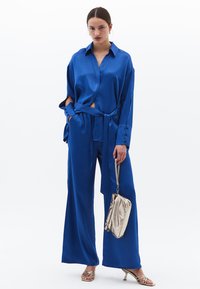 Blusa in satin blu con maniche lunghe e vita legata, abbinata a pantaloni blu a gamba larga. Clutch metallica dorata e sandali con cinturini.