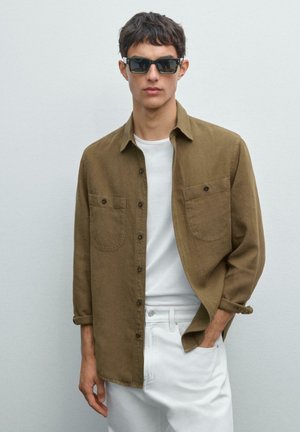Braunes Hemd mit Knöpfen und zwei Brusttaschen, über einem weißen T-Shirt getragen, kombiniert mit weißen Hosen. Das Modell trägt dunkle Sonnenbrillen.