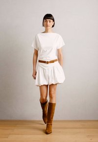 Top blanc à manches courtes, associé à une jupe volumineuse blanche et une ceinture marron. Des bottes en daim marron jusqu'aux genoux complètent le look.