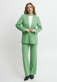 Grön blazer och matchande byxor i ett slätt tyg. Blazern har en insvängd krage och bärs över en vit skjorta. Svarta högklackade skor.