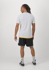 Maglietta atletica bianca con accenti gialli, maniche corte e scollo tondo, abbinata a pantaloni neri con bordi bianchi.