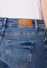 Blue denim jeans featuring a brown leather label on the back waistband. Notable details include back pockets and subtle distressed areas.