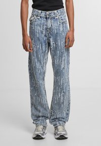 Pantalones vaqueros de denim desgastado en un tono azul claro, con un ajuste holgado, que presentan costuras blancas en contraste y bolsillos frontales. Combinados con zapatillas deportivas grises.