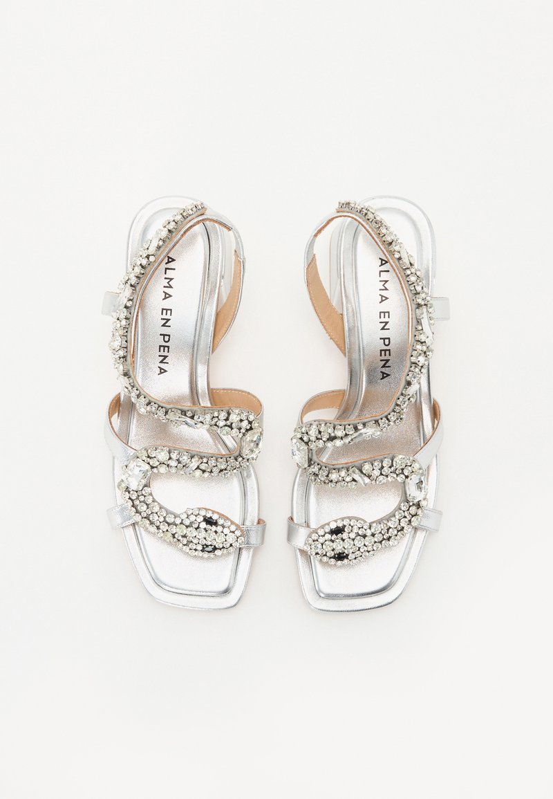 Sandales à talons argentées avec une finition métallique lisse, des brides croisées et ornées de cristaux transparents et d'un motif décoratif.