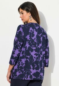 Violetti paita, jossa on tummansinisestä kukkakuviosta, kolmivartiset hihat ja rento istuvuus, valmistettu pehmeästä, rakenteellisesta materiaalista.