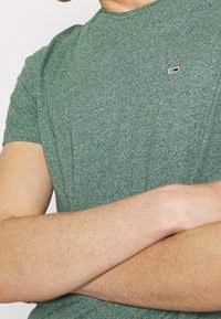 Osoba w zielonej koszuli z krótkim rękawem z małym logo, z założonymi na klatkę piersiową rękami, na neutralnym tle.