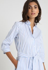 Kvinde iført en lyseblå og hvidt lodretstribet skjortekjole med trekvartlange ærmer og bindebånd i taljen, stående foran en ensfarvet baggrund.