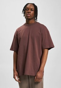 Camiseta de gran tamaño en color marrón, hecha de algodón suave, con cuello redondo, mangas cortas y bajo recto, adornada con una pequeña etiqueta de logo en el lateral.