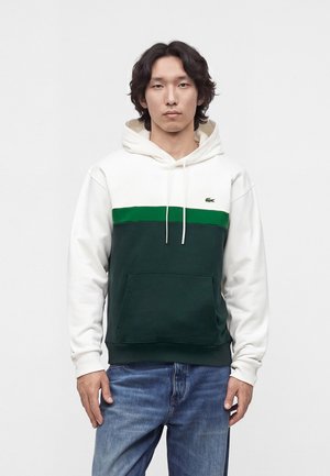 Jonge man met halflang haar die een witte, groene en donkergroene colorblock-hoodie en blauwe spijkerbroek draagt, staand tegen een effen achtergrond.