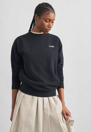 Woman with braided hair wearing a black Ellesse sweatshirt and beige pleated skirt, looking down to her left against a plain background.