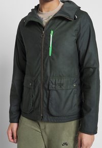 Dark green jacket with a hood, featuring a front zipper, two large flap pockets, and a small zippered pocket with a green accent detail.