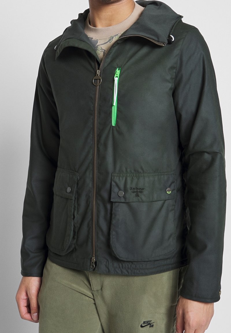 Dark green jacket with a hood, featuring a front zipper, two large flap pockets, and a small zippered pocket with a green accent detail.