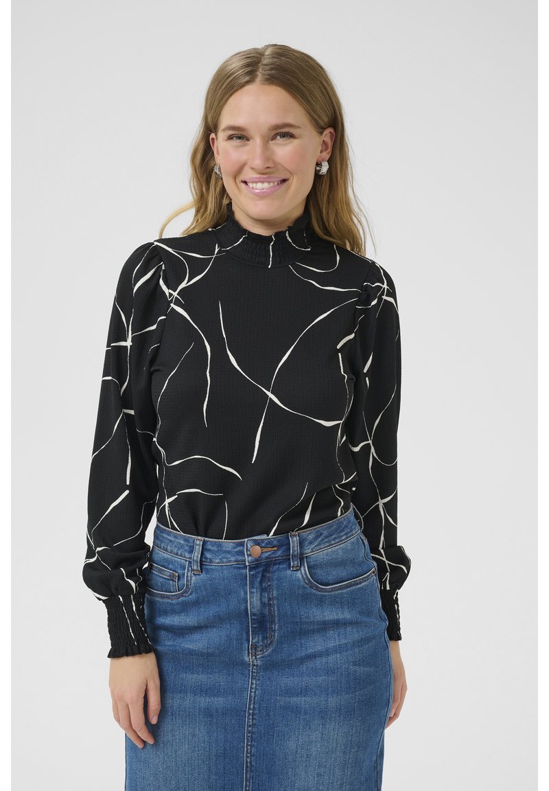 Sort langærmet top med hvide abstrakte linjer, høj hals og plisserede ærmer, parret med en denimskørt med en tætsiddende talje.