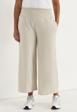 Woman wearing loose, cream wide-leg pants with a gathered elastic waistband and white athletic shoes, standing against a plain background.