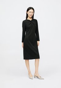 Crna haljina dugih rukava s dizajnom uvijenog prednjeg dijela, do koljena, izrađena od glatkog materijala. Nosena s svijetlim cipelama na špic.