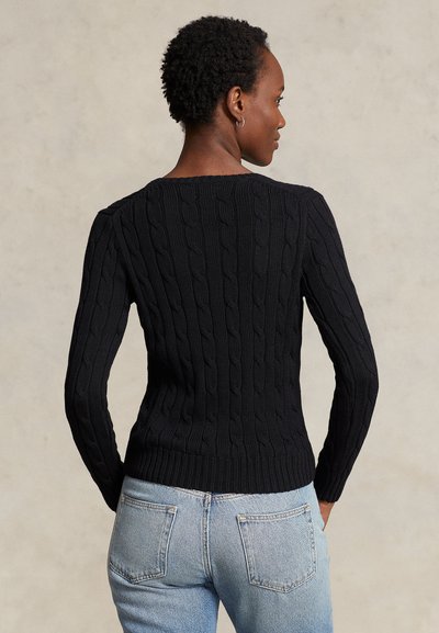 Ženska oblečena v črni pulover z vozlanim vzorcem in svetlo modre jeans hlače, prikazana od zadaj na enostavnem ozadju.