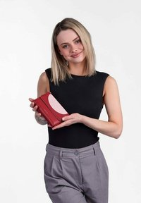 Red leather wallet with a curved pink accent, smooth texture, and logo detail, held by a person in a black top and gray pants.