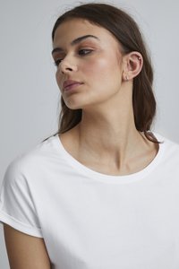 Bílý bavlněný tričko s ohrnutými rukávy, kulatým výstřihem a hladkou texturou. Modelka má minimální šperky a přirozený make-up.