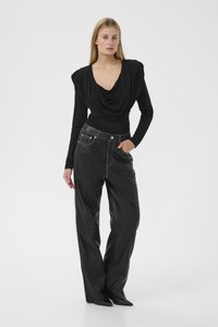 Blond kvinne står med hånd i lommen iført svart drapert topp med polstrede skuldre og svarte høytlivs skinnbukser med hvit søm.