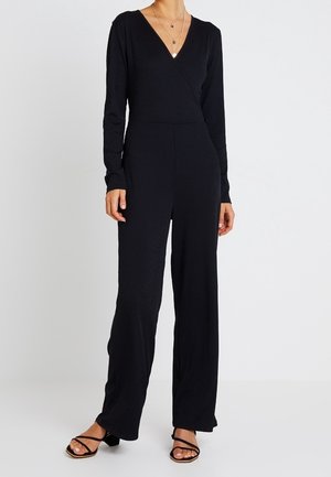 Czarny prążkowany kombinezon z długimi rękawami i dekoltem w serek. Luźny, szeroki krój nogawek. Uzupełniony warstwowymi naszyjnikami i sandałami na paski.