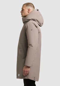 Parka beige chiaro con cappuccio, maniche lunghe e tasche laterali. Presenta una finitura liscia e opaca e un'etichetta del marchio ben visibile all'orlo.