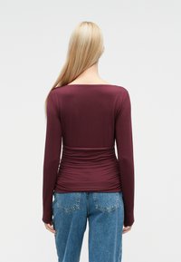 Burgundy long-sleeve top with a fitted waist and ruched detailing, paired with high-waisted blue denim jeans. Smooth texture, simple design.
